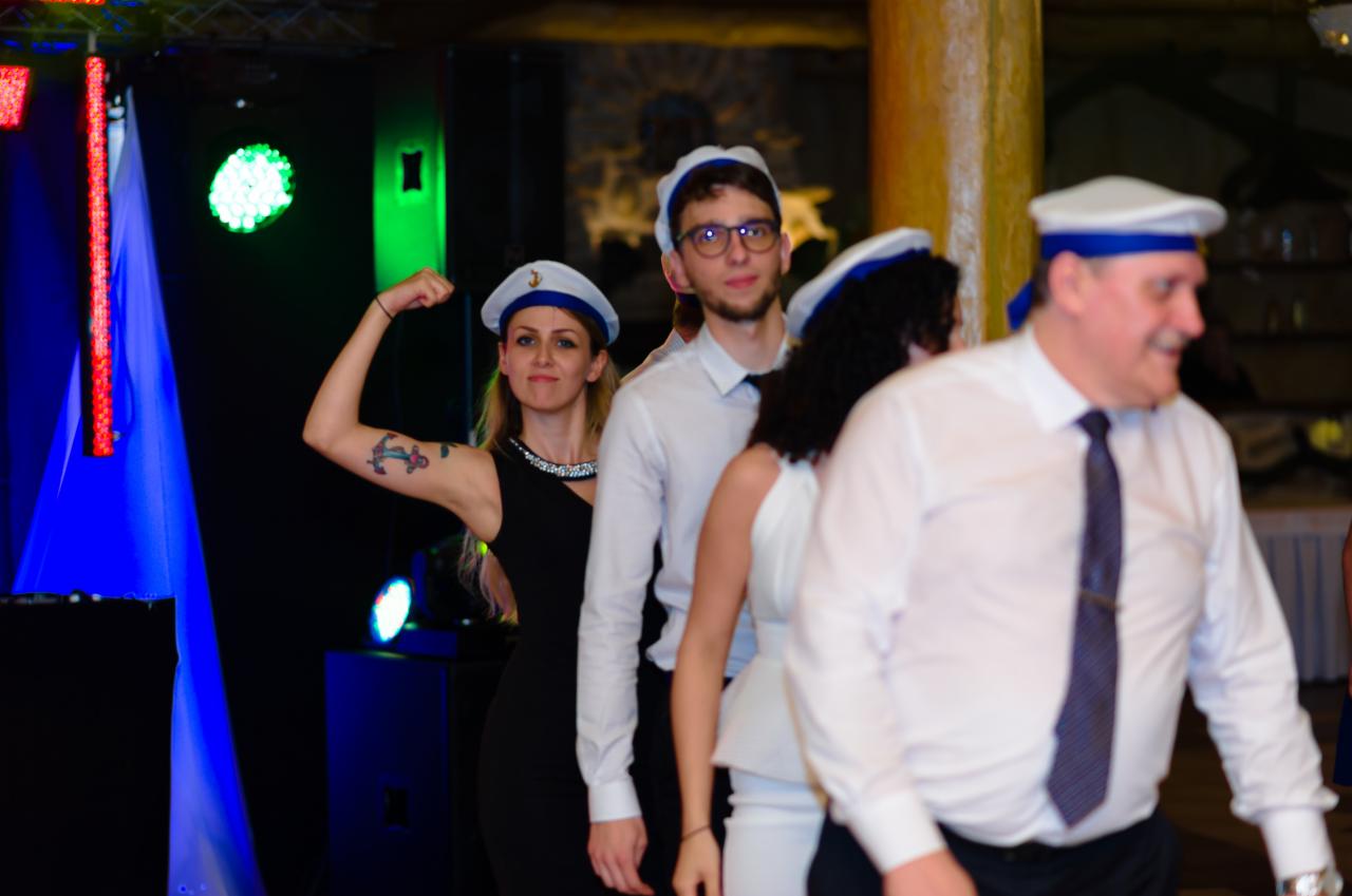 Grupa osób w marynarskich czapkach na imprezie, kobieta z tatuażem kotwicy na ramieniu prezentuje muskuły, oświetlenie sceniczne w tle.