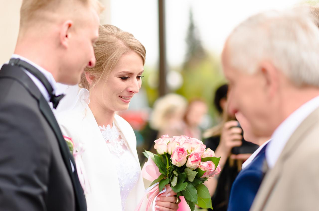Uśmiechnięta panna młoda w białej sukni i bolerku trzyma bukiet różowych róż, obok pan młody w smokingu i starszy mężczyzna w garniturze, w tle rozmyte sylwetki gości.