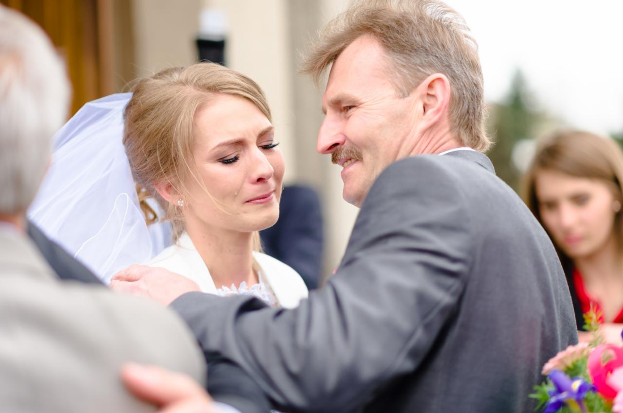 Wzruszona panna młoda w welonie, ocierająca łzy podczas błogosławieństwa przez ojca z wąsami, w tle zamazana postać kobiety.