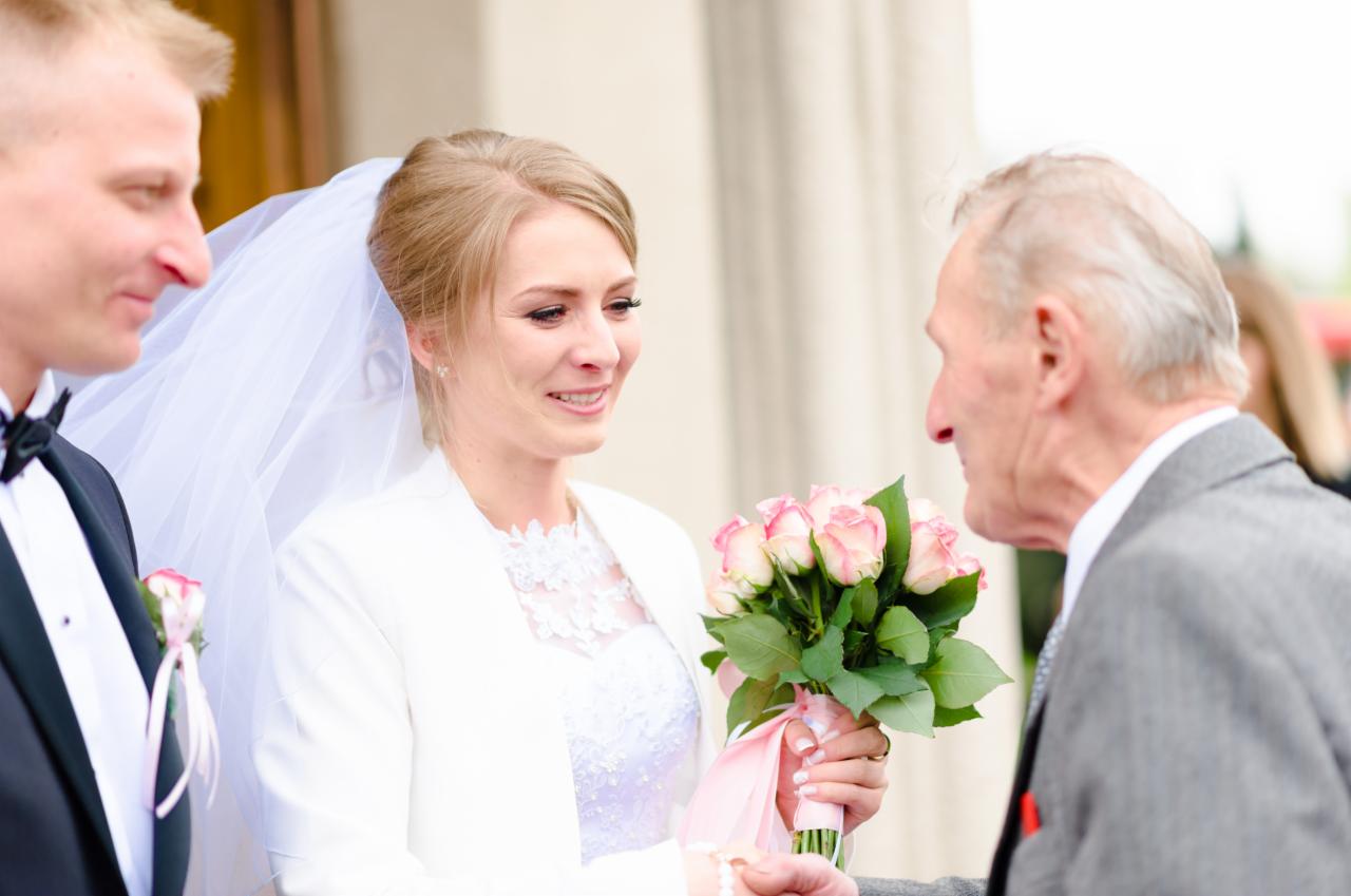Wzruszona panna młoda w białym żakiecie i welonie, trzyma bukiet różowych róż, spogląda na starszego mężczyznę w szarym garniturze, obok stoi pan młody w smokingu.