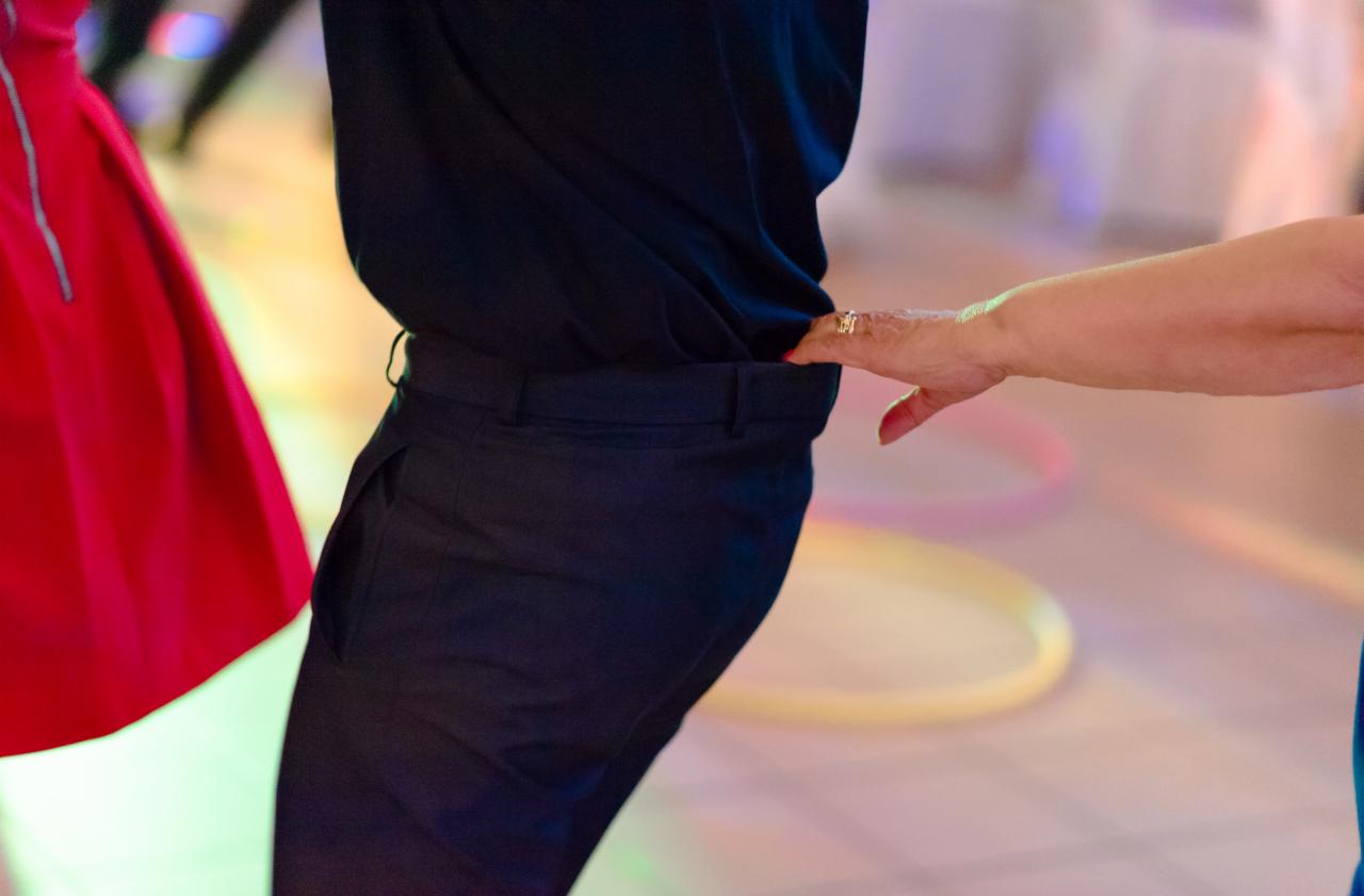 Zbliżenie na parę podczas tańca: dłoń kobiety z pierścionkiem dotyka talii mężczyzny w ciemnych spodniach, w tle fragment czerwonej sukienki i rozmyte, kolorowe światła.