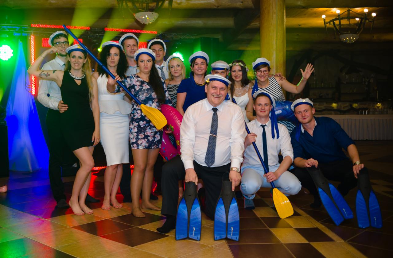 Grupa osób w marynarskich czapkach z wiosłami i płetwami w sali z parkietem, oświetlona kolorowymi światłami.