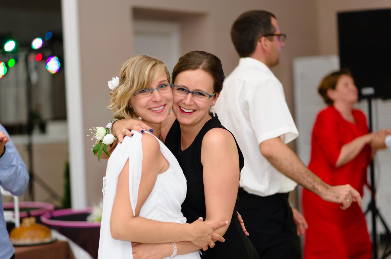 Panna młoda w białej sukni i druhna w czarnej sukience obejmują się na sali weselnej podczas zabawy, w tle widoczni inni goście i dekoracje.