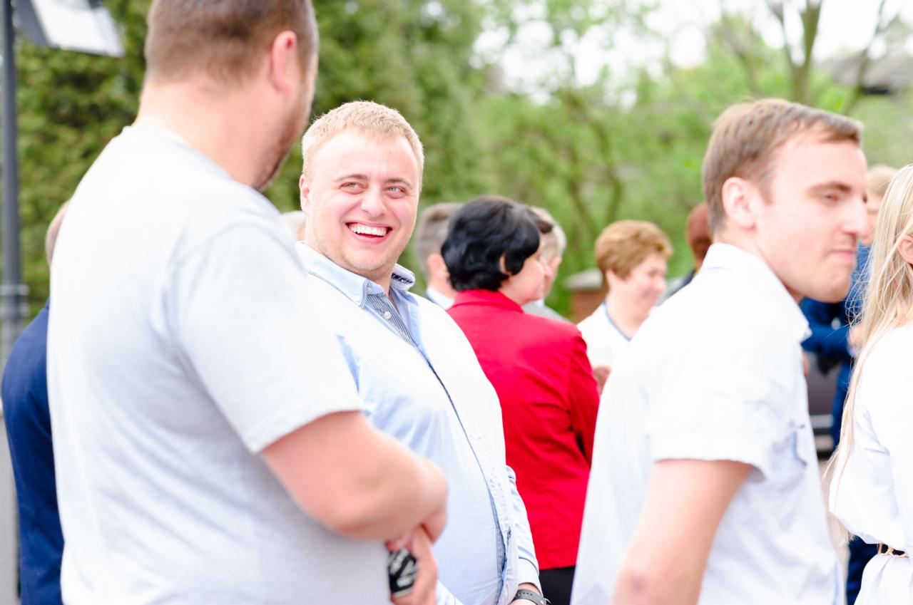 Uśmiechnięty mężczyzna w błękitnej koszuli, otoczony przez grupę osób podczas wydarzenia na zewnątrz, z zielenią w tle.