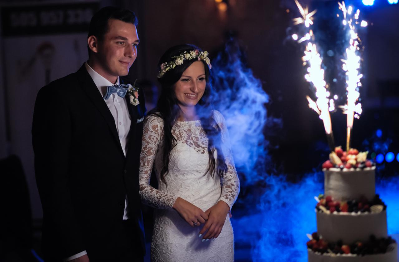 Uśmiechnięta para młoda obok tortu weselnego z zimnymi ogniami na tle niebieskiego dymu.
