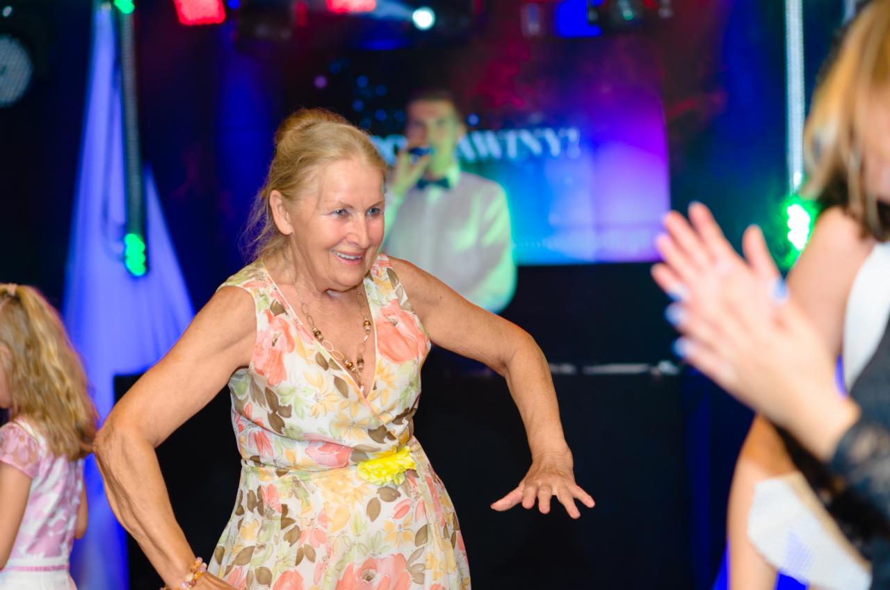 Uśmiechnięta starsza kobieta w kwiecistej sukience tańczy na parkiecie podczas imprezy z DJ-em w tle, oświetlona kolorowymi światłami.