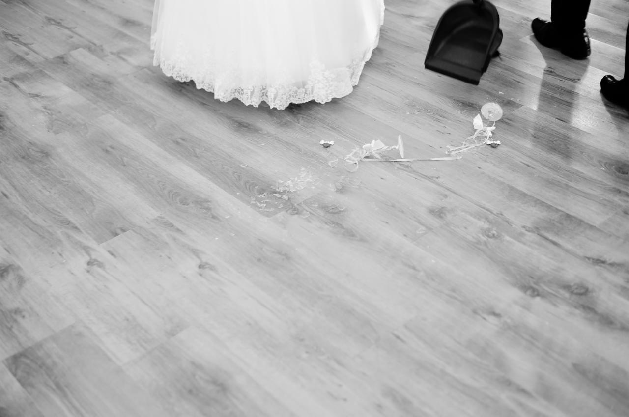 Rozbite szkło i fragmenty dekoracji po wypadku ze szklanką podczas uroczystości ślubnej w Jastrzębiu-Zdroju, obok widoczny fragment sukni ślubnej i zmiotka.