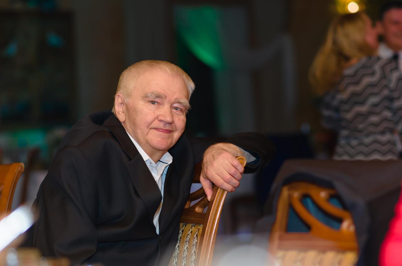 Portret starszego mężczyzny w ciemnym garniturze, siedzącego na krześle z drewnianym oparciem, w tle para w nieostrym ujęciu.