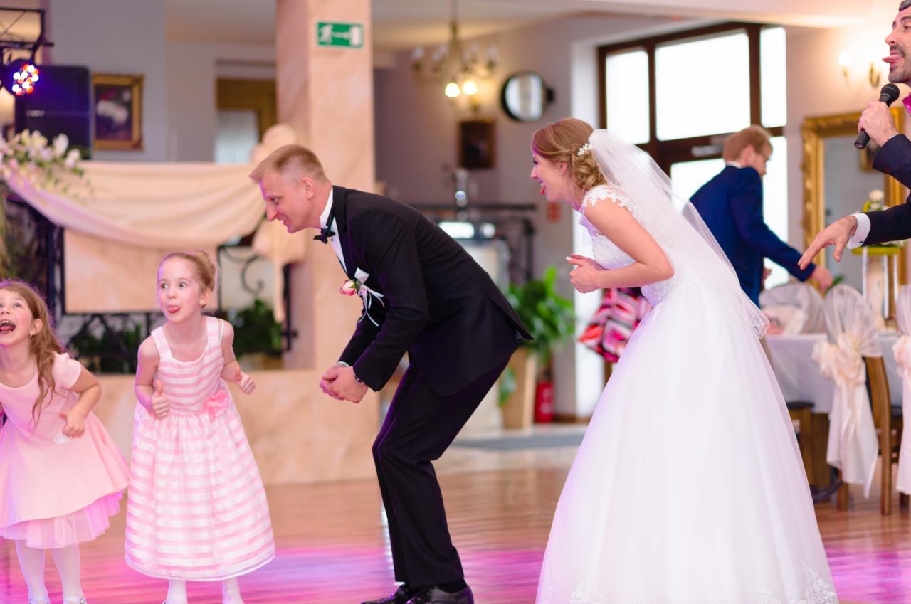 Para młoda w trakcie zabawy weselnej z dziećmi, wodzirej z mikrofonem w tle, radosne miny i wyciągnięte języki.