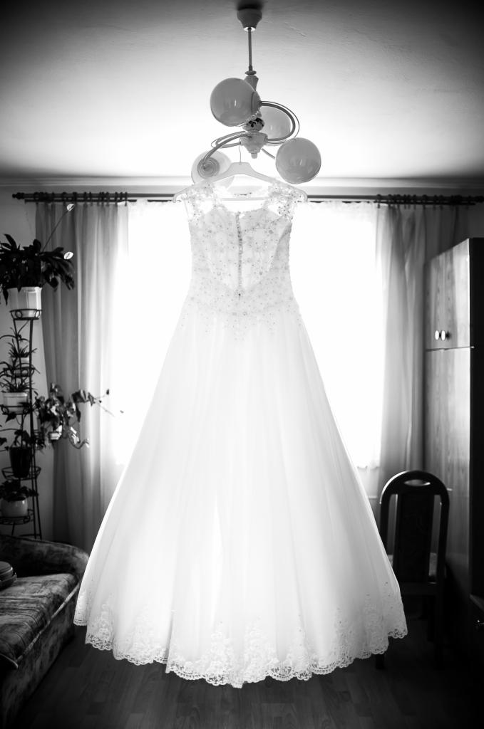 Czarno-białe zdjęcie sukni ślubnej wiszącej na wieszaku pod lampą w pokoju z oknem w tle, widoczny fragment sofy i szafy.