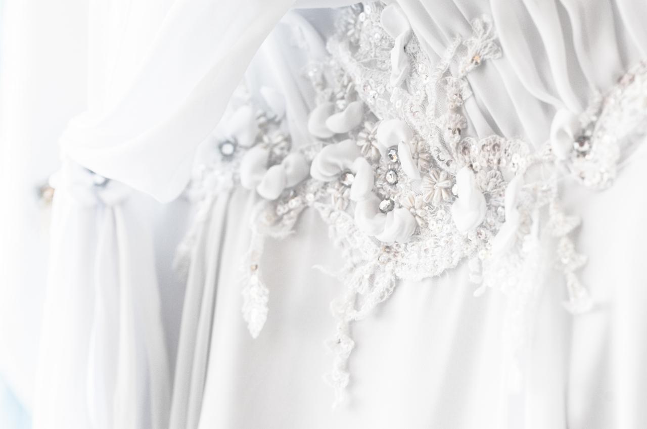 Detal koronkowego zdobienia białej sukni z aplikacjami z materiału i błyszczącymi kamieniami, delikatnie oświetlona naturalnym światłem.