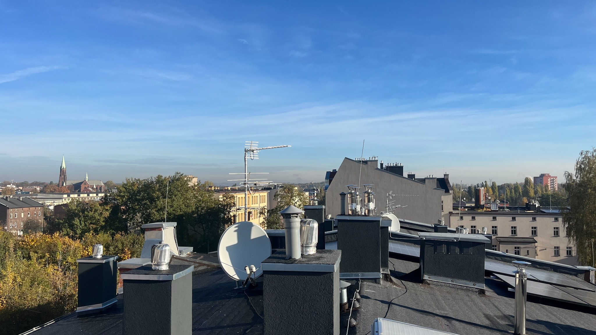 Anteny satelitarne i telewizyjne na dachu budynku z widokiem na miasto w tle, w słoneczny dzień. Widoczne kominy i elementy instalacji.