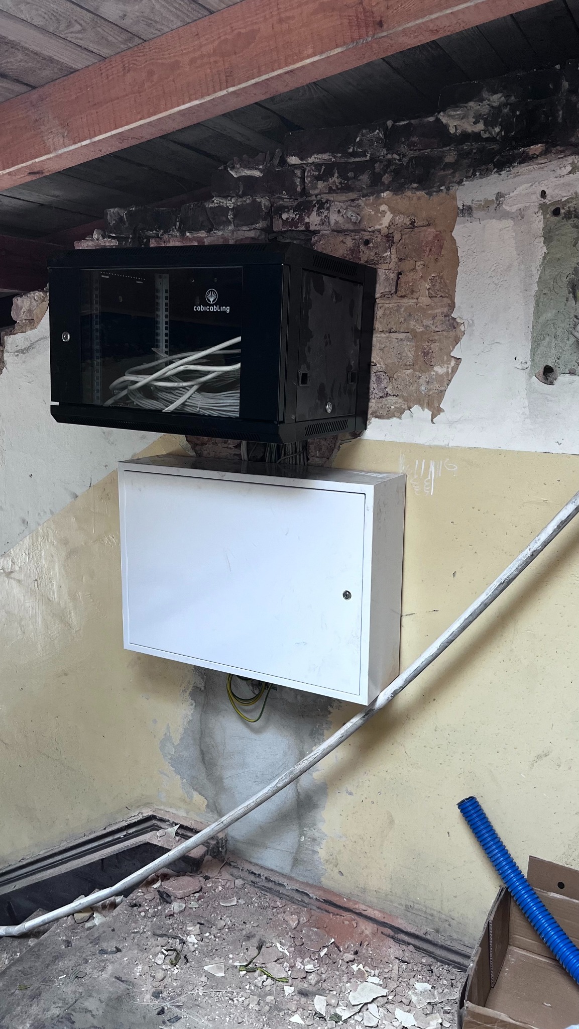 Czarna szafa rack z okablowaniem i biała skrzynka na ścianie z odsłoniętą cegłą i elementami instalacji. Widoczne przewody i rury instalacyjne.