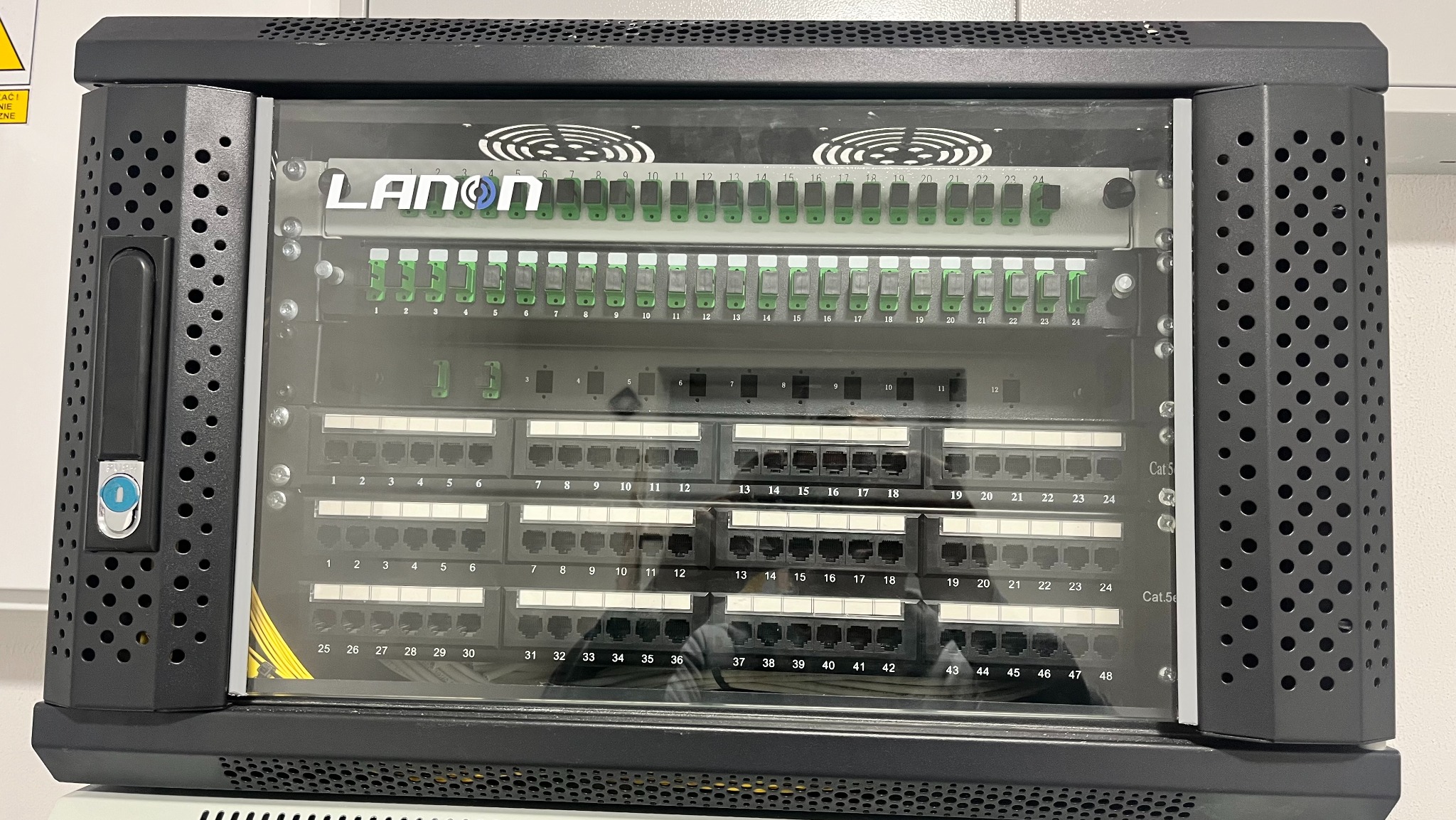 Panel krosowniczy LANON w szafie rack, z widocznymi portami RJ45 i numeracją. Element instalacji telekomunikacyjnej, z wentylatorami u góry i perforowaną obudową.