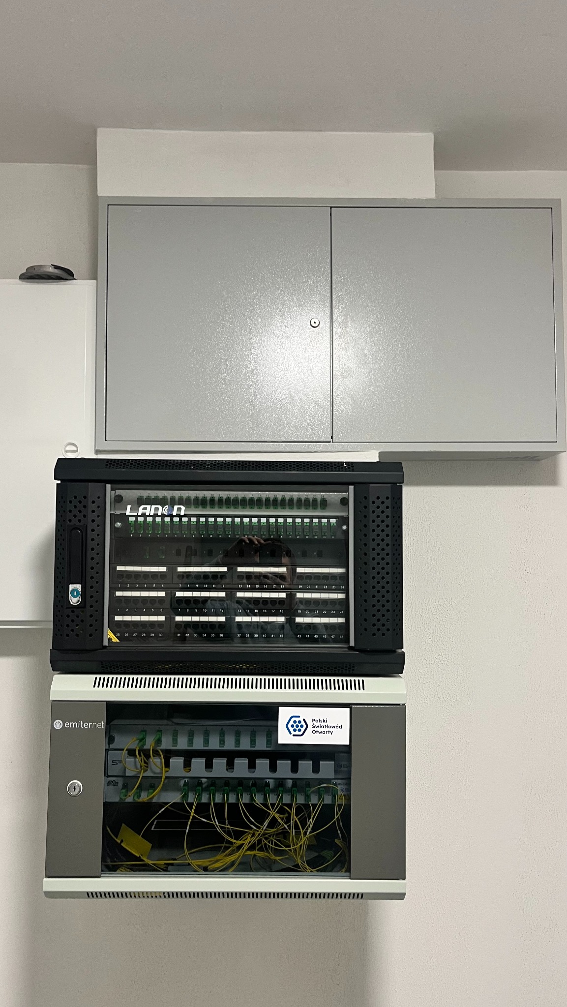 Szafa telekomunikacyjna LANDA i emiter.net z patch panelem i światłowodami, zamontowana na ścianie w pomieszczeniu technicznym. Widoczne kable i oznaczenia portów.