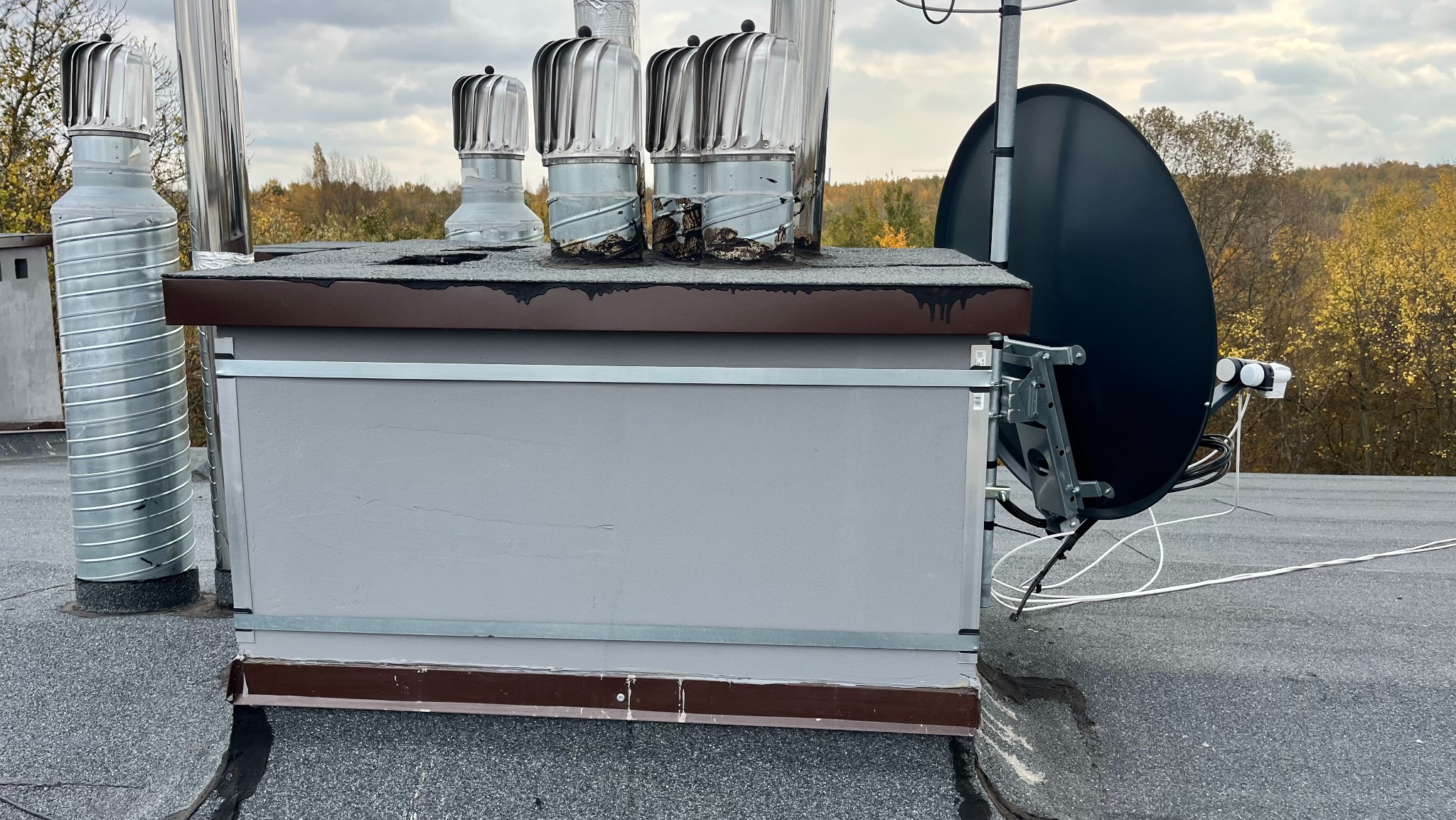 Antena satelitarna zamontowana na dachu budynku, obok kominów wentylacyjnych. Widoczne przewody antenowe i elementy mocujące. Tło stanowią drzewa i pochmurne niebo.