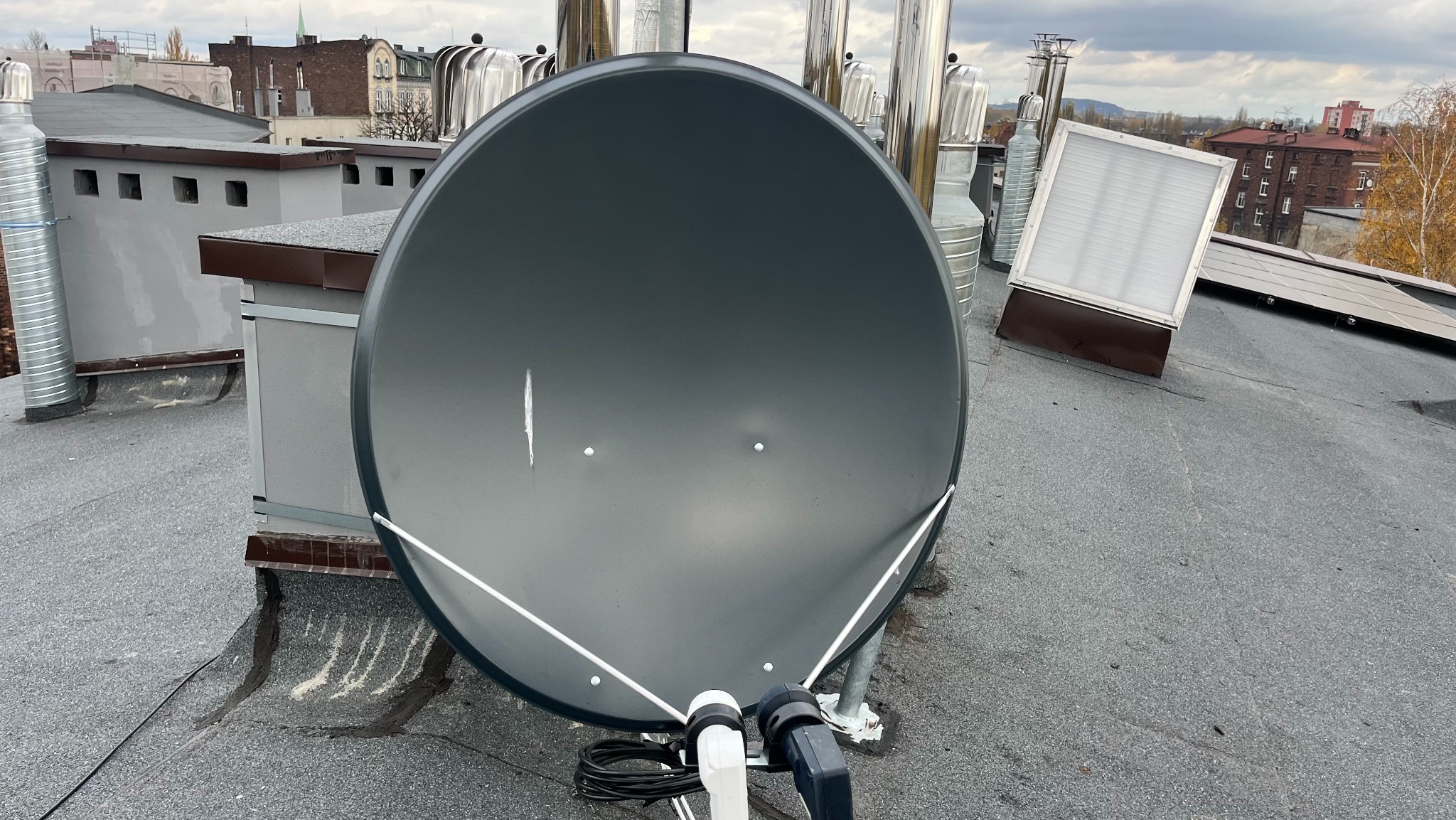 Antena satelitarna zamontowana na dachu budynku z widocznymi elementami instalacji i otoczeniem miejskim w tle, uchwycona z bliskiej perspektywy.