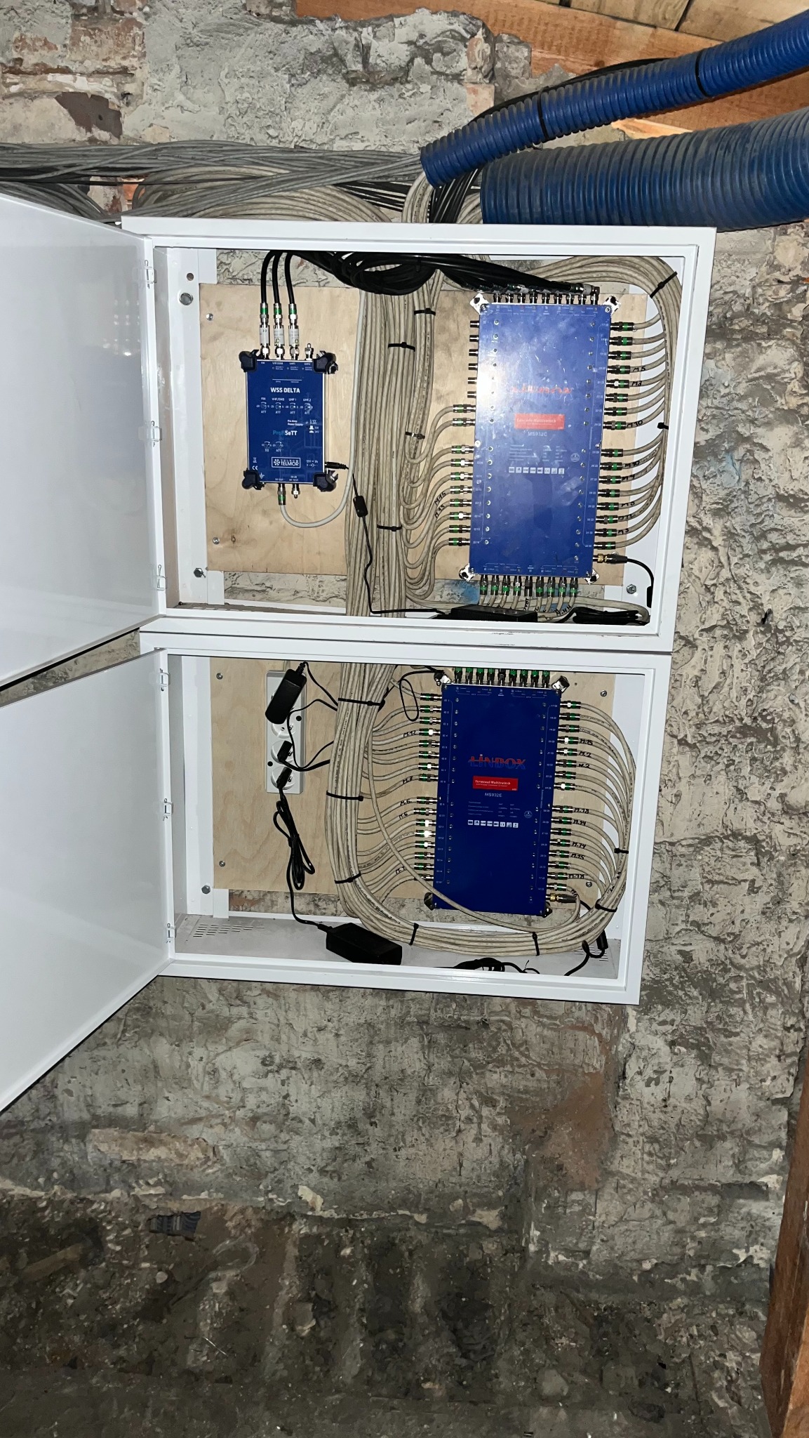 Dwa białe panele z rozdzielaczami sygnału i kablami koncentrycznymi, zamontowane na ścianie z cegły. Widoczne zasilacze i kable w uporządkowanym układzie.