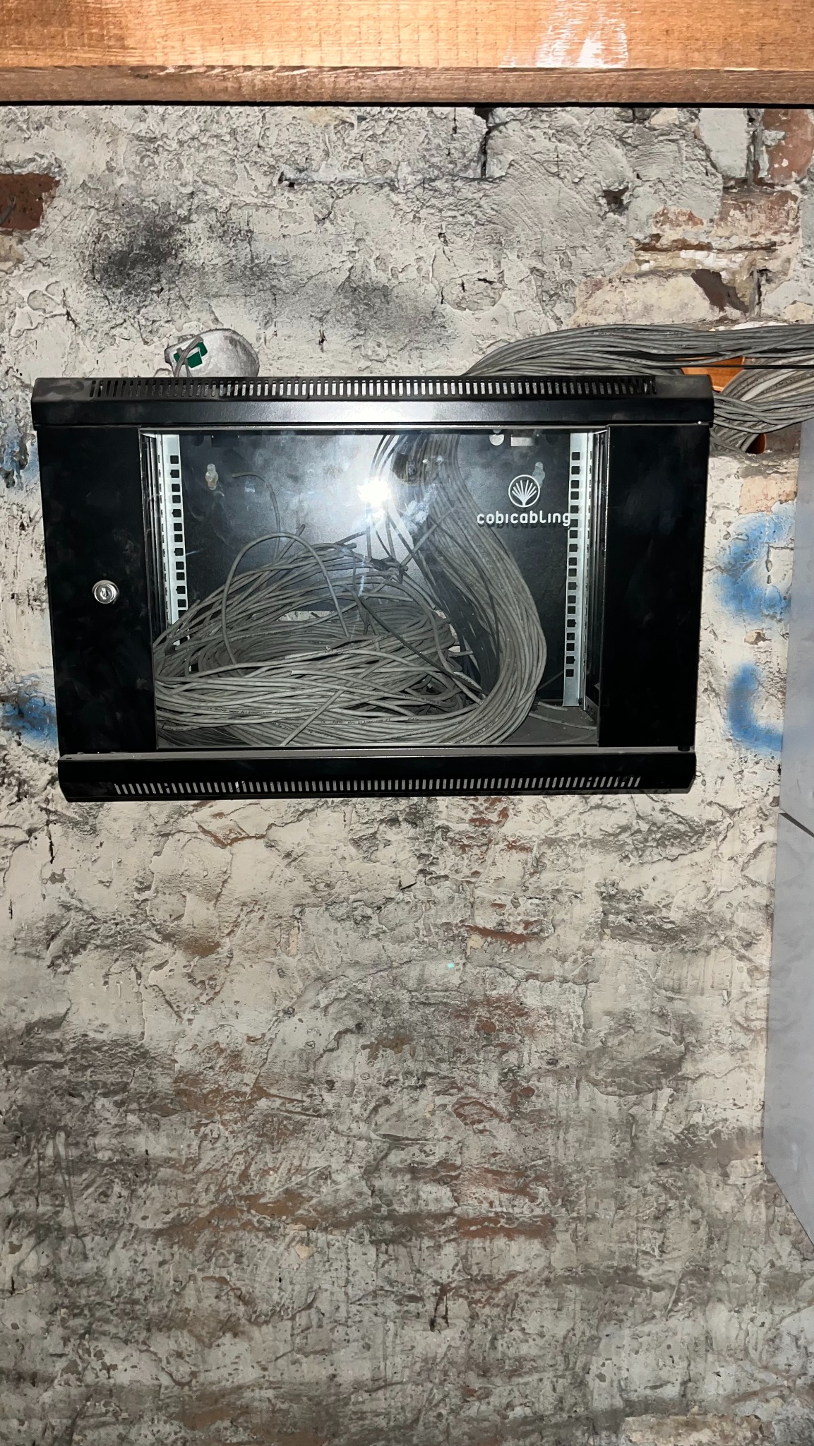 Czarna szafka rack z transparentnymi drzwiami, wypełniona szarymi kablami, zamontowana na starej, pomalowanej ścianie z cegły. Widoczne logo 'cobicabling'.