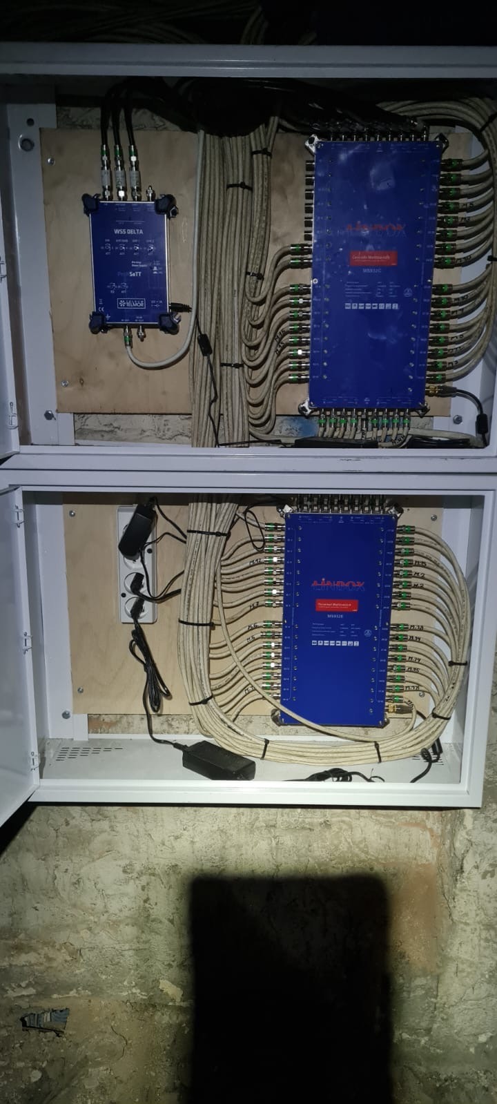 Dwa panele rozdzielcze w białych skrzynkach, z licznymi kablami koncentrycznymi podłączonymi do niebieskich modułów. Widoczne zasilacze i okablowanie.