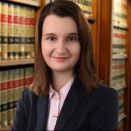 Portret uśmiechniętej kobiety w ciemnej marynarce i różowej koszuli, na tle regałów z książkami w gabinecie.