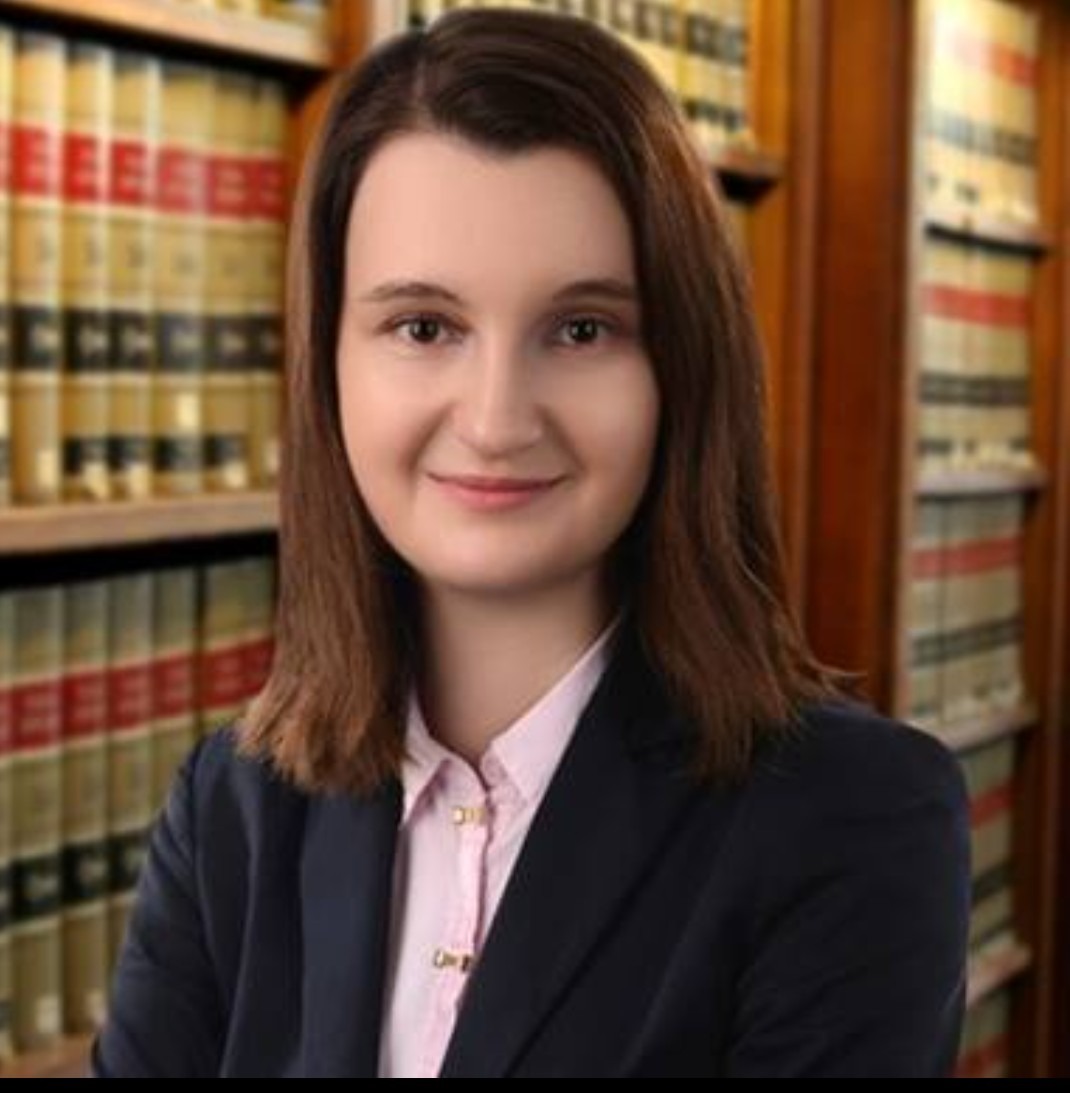 Portret uśmiechniętej kobiety w ciemnej marynarce i różowej koszuli, na tle regałów z książkami w gabinecie.