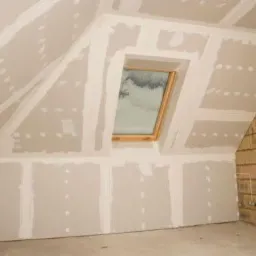 Poddasze w trakcie wykańczania: ściany i sufit pokryte płytami gipsowo-kartonowymi, widoczne łączenia przed szpachlowaniem, okno dachowe z drewnianą ramą, fragment ściany z cegły.