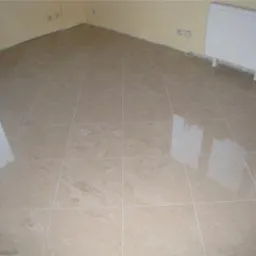 Jasne, beżowe płytki podłogowe z wyraźnym połyskiem, ułożone w regularną siatkę, odbijające światło okna w świeżo wyremontowanym pokoju z żółtymi ścianami i białym kaloryferem.