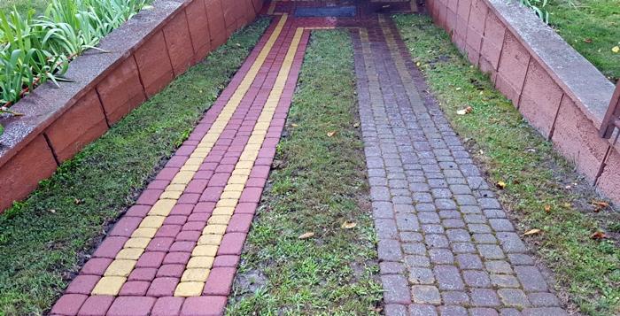 Porównanie kostki brukowej przed i po czyszczeniu: po lewej stronie kostka czerwono-żółta, po prawej szara, widoczna różnica w kolorze i czystości, trawa pomiędzy kostką i przy murku.