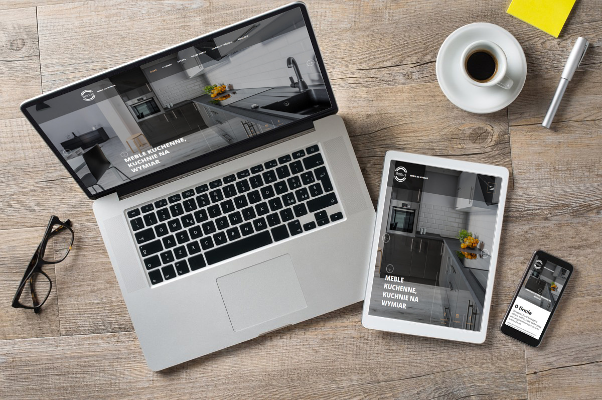 Laptop, tablet i smartfon wyświetlają tę samą stronę o meblach kuchennych na wymiar, obok filiżanka kawy, notatnik, długopis i okulary na drewnianym blacie.