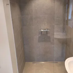 Nowoczesna kabina prysznicowa z szarymi płytkami imitującymi kamień, chromowaną armaturą i odpływem liniowym w podłodze, oddzielona od reszty łazienki szklaną ścianką.