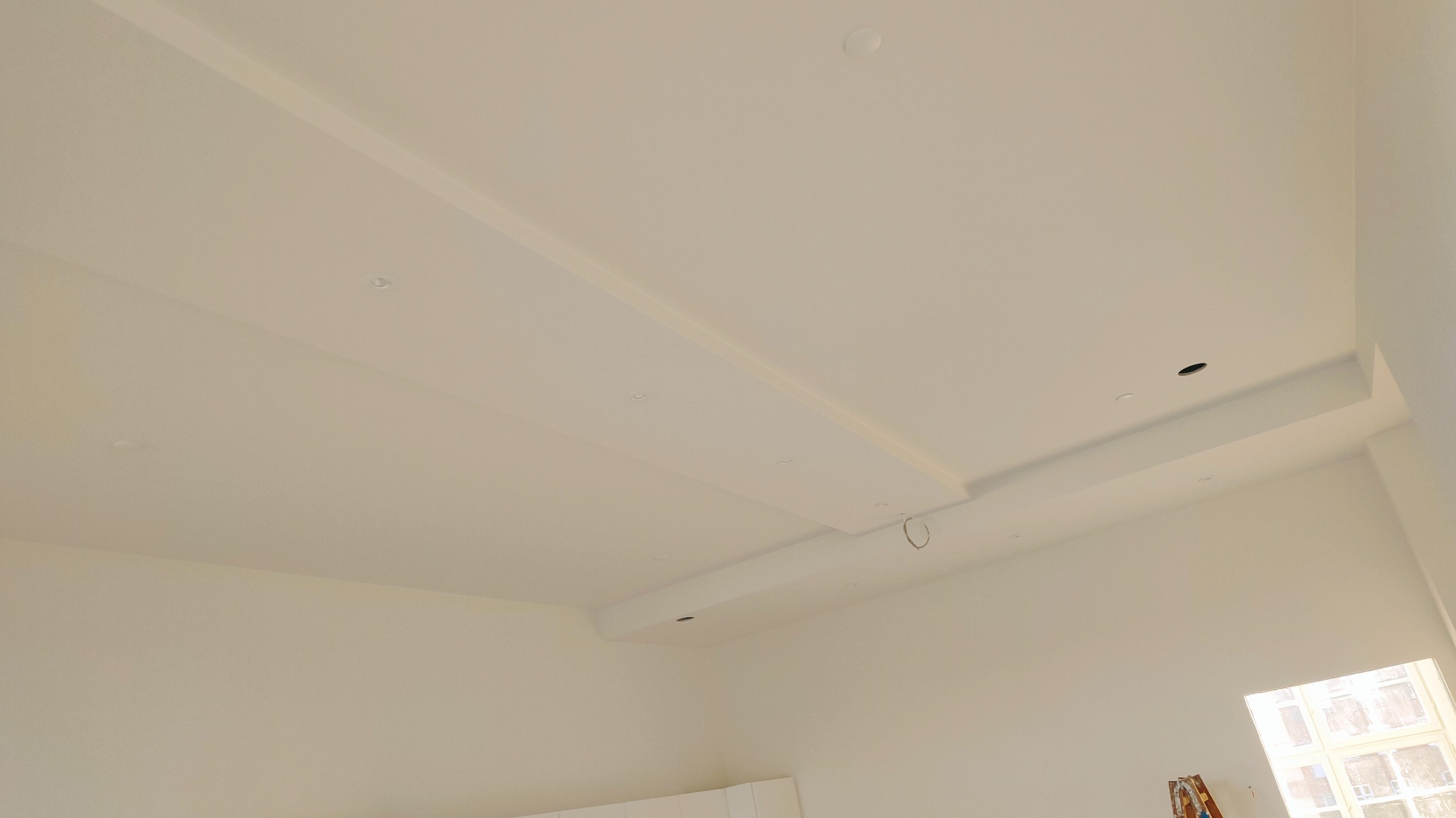 Białe wnętrze w trakcie wykańczania, widoczny sufit z podwieszanymi elementami i otworami na oświetlenie, ściany pomalowane na biało, fragment drabiny i okna w dolnej części kadru.