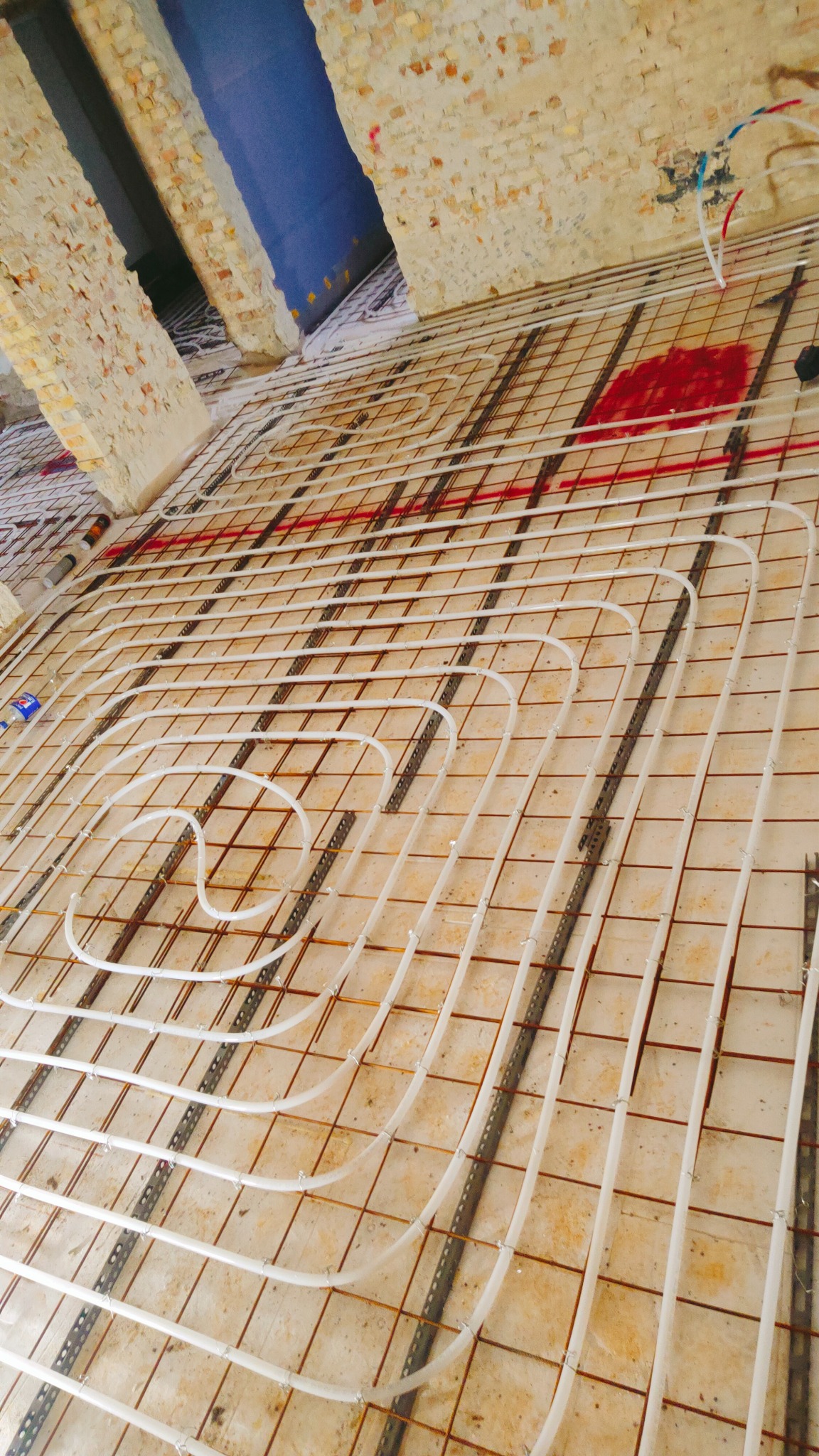 Układ ogrzewania podłogowego w trakcie instalacji, rury grzewcze ułożone na metalowej siatce, widoczne fragmenty surowych ścian z cegły i instalacje hydrauliczne.
