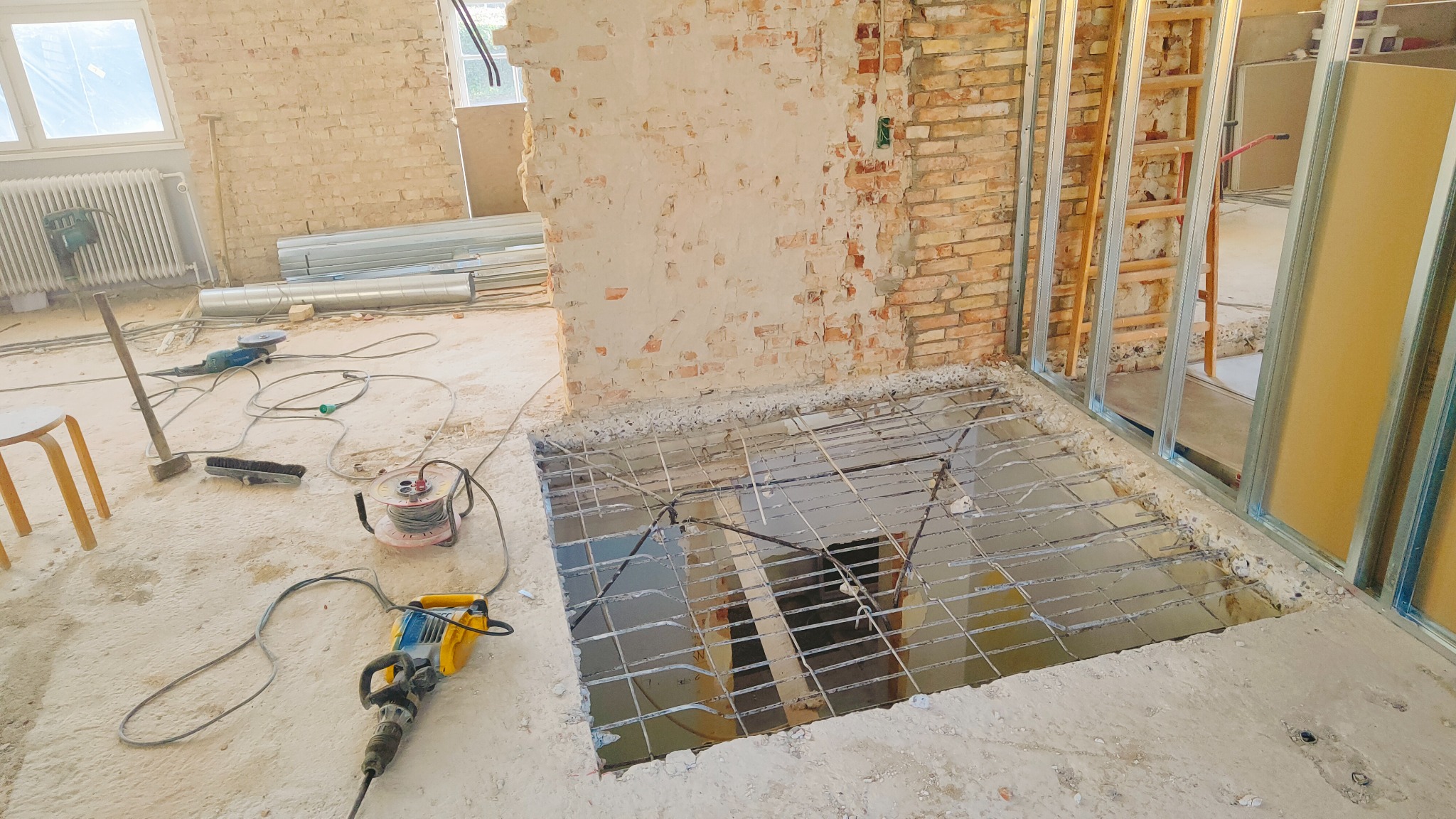 Przygotowanie podłoża pod podłogę: siatka zbrojeniowa ułożona w wyciętym otworze w posadzce, widoczne metalowe profile konstrukcji ścianki działowej, fragment muru z cegły i narzędzia budowlane...