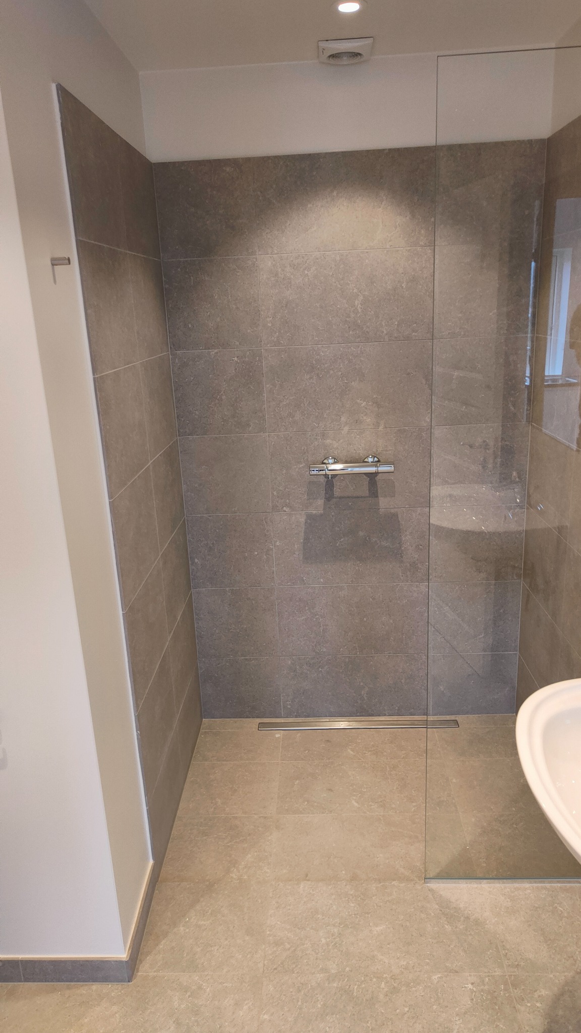 Nowoczesna kabina prysznicowa z szarymi płytkami imitującymi kamień, chromowaną armaturą i odpływem liniowym w podłodze, oddzielona od reszty łazienki szklaną ścianką.