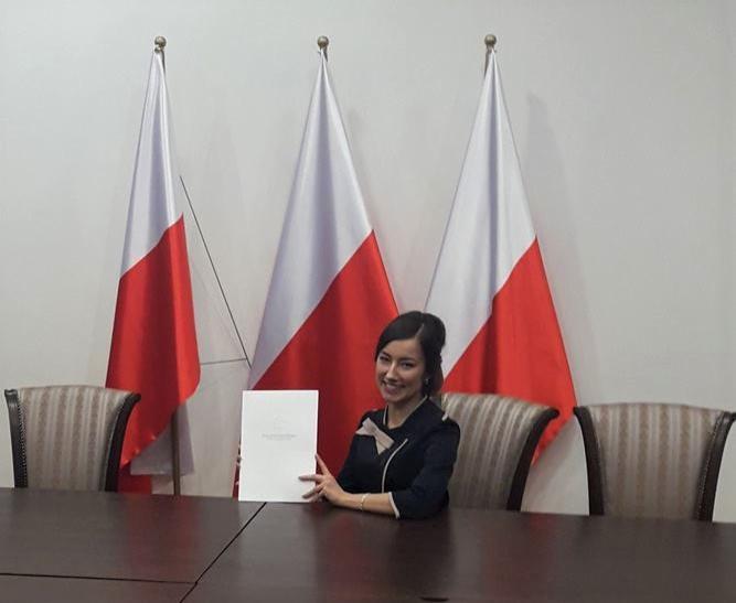 Kobieta w ciemnej marynarce z dekoracyjną kokardą siedzi za ciemnym stołem i trzyma w rękach dokument na tle trzech biało-czerwonych flag.