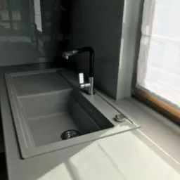 Szary, dwukomorowy zlewozmywak z czarną baterią, zamontowany w blacie przy oknie z roletą rzymską.