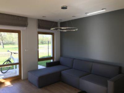 Nowoczesny salon z szarą sofą narożną, okrągłą lampą wiszącą i oknami balkonowymi z roletami rzymskimi; rower widoczny za oknem.