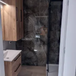 Nowoczesna łazienka z umywalką w szafce z drewnianym fornirem, kabiną prysznicową z czarnymi profilami i marmurowymi płytkami na ścianach.