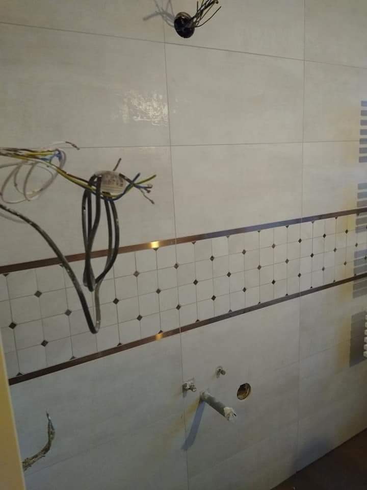 Ściana w trakcie prac wykończeniowych z widocznymi kablami elektrycznymi, rurami hydraulicznymi i płytkami ceramicznymi z dekoracyjnym pasem. Widoczne otwory w ścianie przygotowane pod osprzęt.