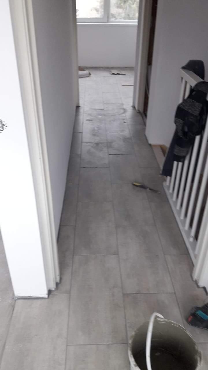 Korytarz w trakcie układania szarych płytek podłogowych, widoczny biały kubeł i narzędzia, fragment białej balustrady schodów, drzwi po lewej stronie z widocznym fragmentem wzoru na skrzydle.