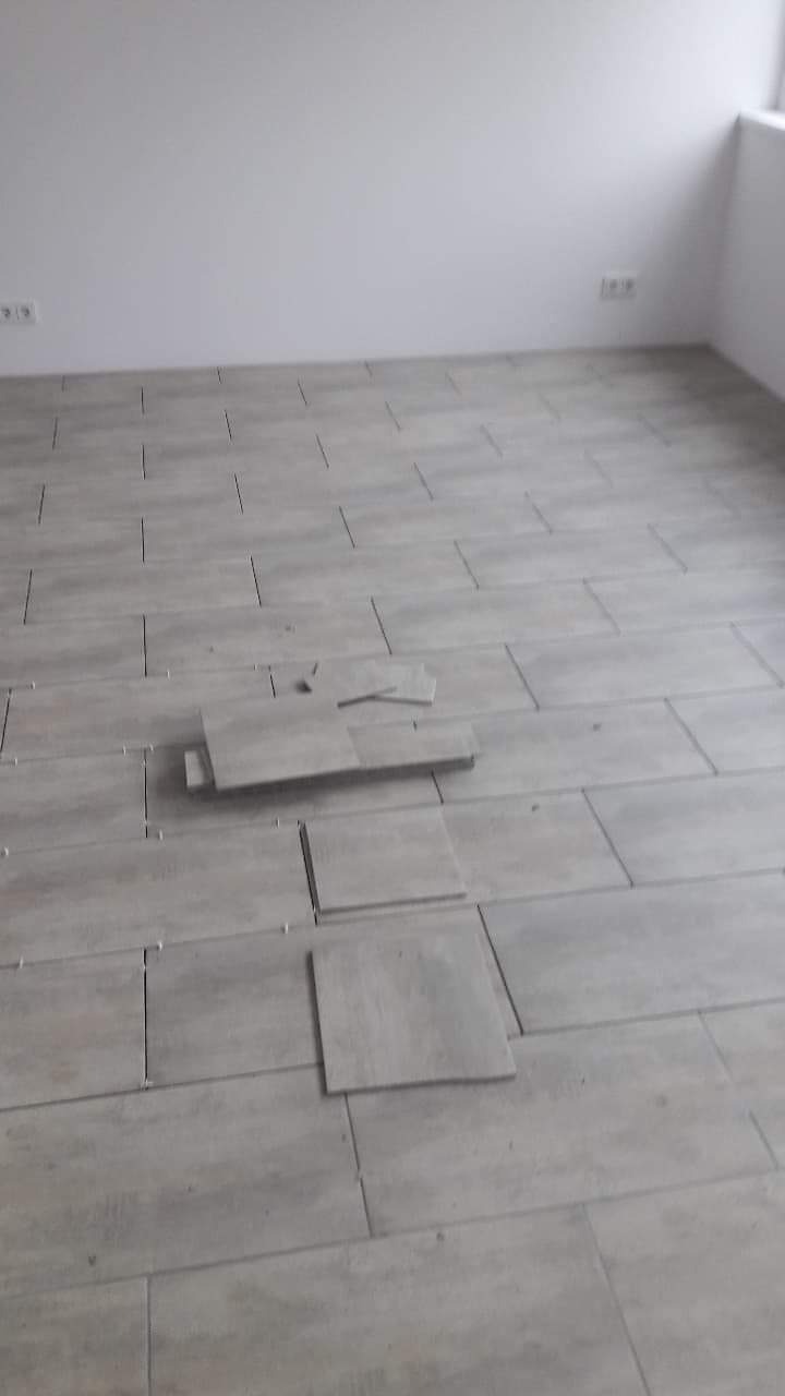 Układanie szarych płytek podłogowych w prostokątnym formacie, widoczne nierówności i kilka płytek ułożonych obok siebie, białe ściany w tle.