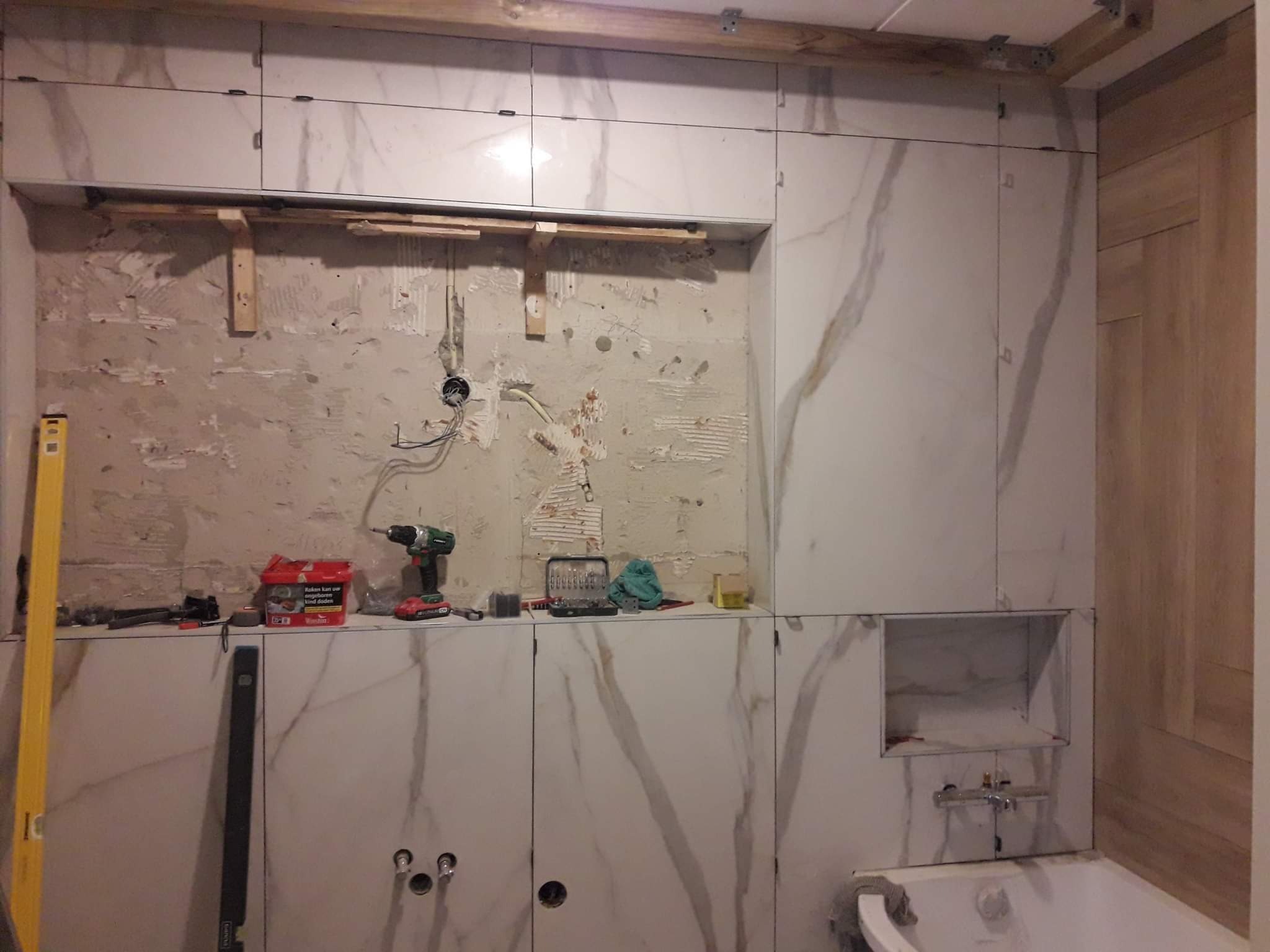 Remont łazienki w trakcie: częściowo ułożone płytki imitujące marmur na ścianie, widoczny fragment surowej ściany pod przyszłą zabudowę, narzędzia pracy i poziomica, oraz fragment wanny.