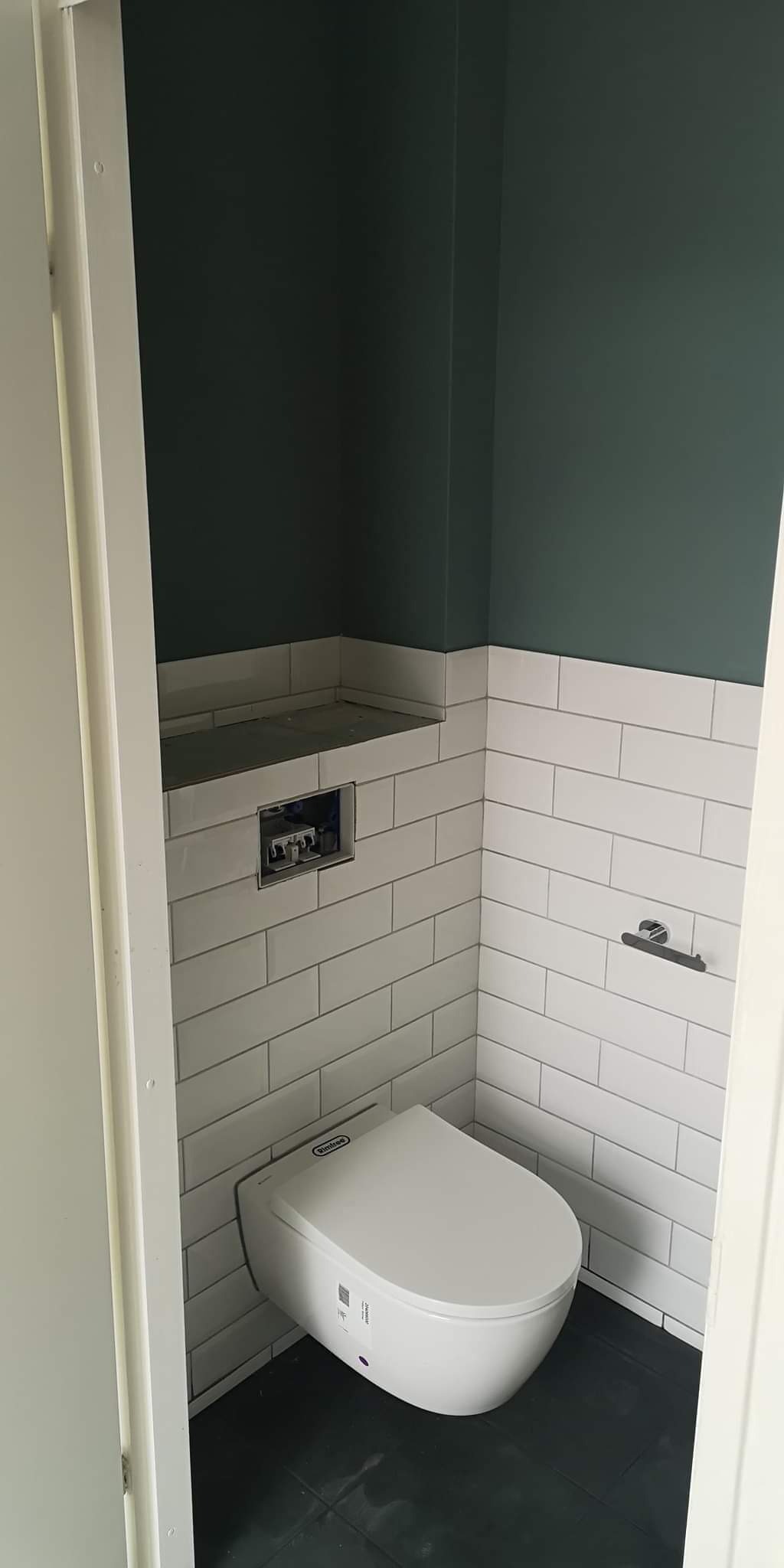 Nowoczesna toaleta podwieszana z ukrytą spłuczką, białe kafelki w stylu cegiełki do połowy ściany, górna część ściany pomalowana na ciemny zielony kolor, ciemna podłoga.