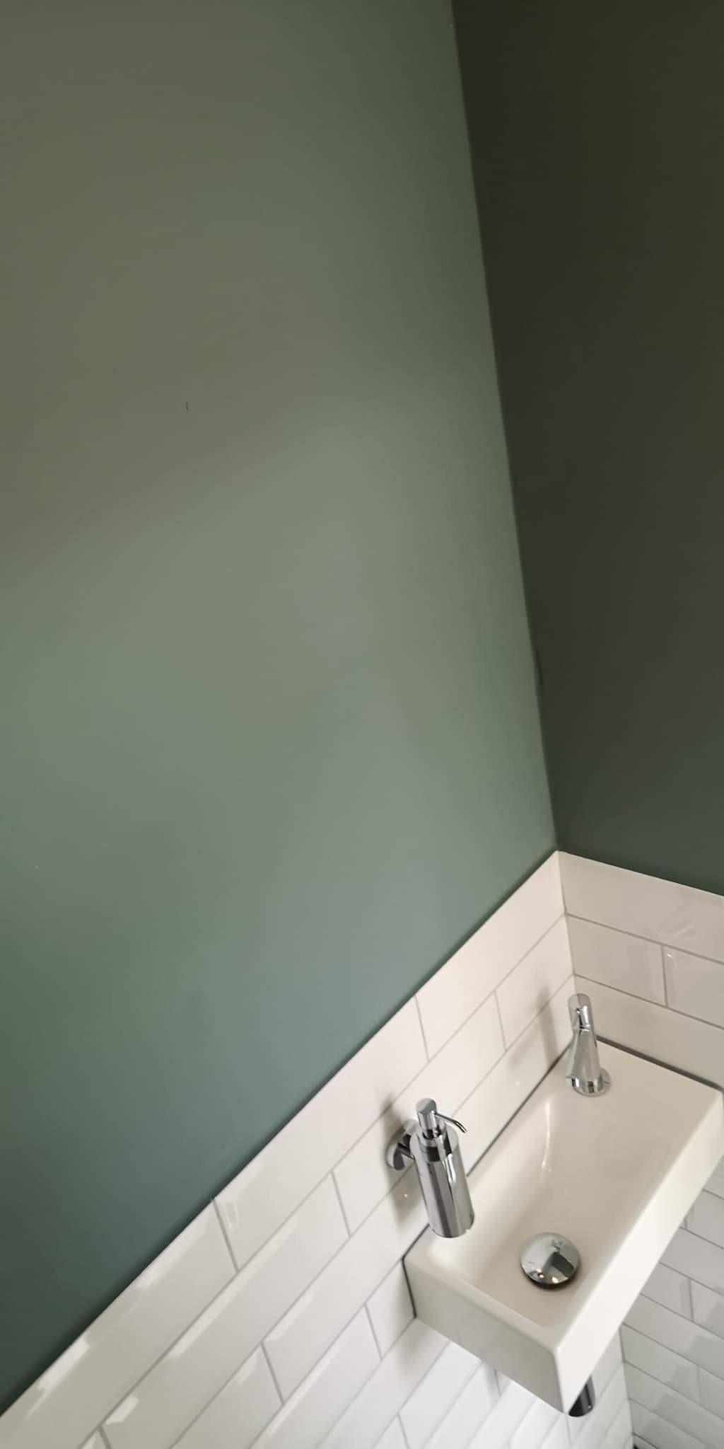 Mała, prostokątna umywalka wbudowana w białe, prostokątne płytki, z zielonymi ścianami w tle, widoczne chromowane baterie i dozownik mydła.