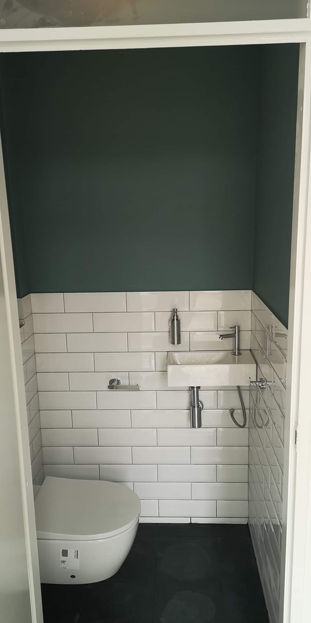 Mała łazienka z toaletą wiszącą, prostokątną umywalką i białymi płytkami metra do połowy ściany, reszta ściany pomalowana na ciemnozielono.