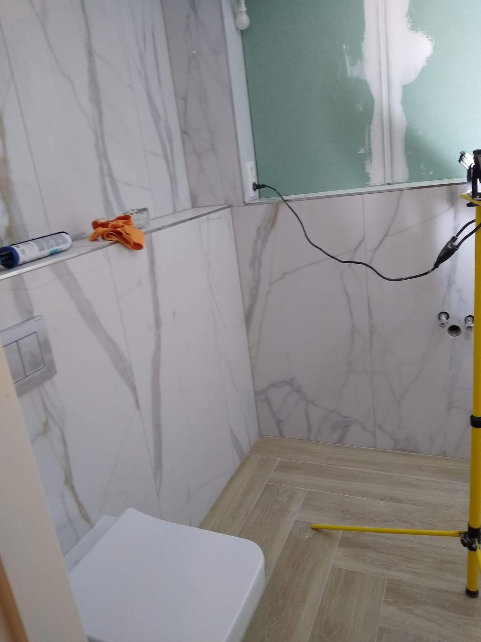 Łazienka w trakcie remontu: białe marmurowe płytki na ścianach, częściowo położona płyta karton-gips, drewnopodobna podłoga ułożona w jodełkę, widoczny fragment toalety, narzędzia i lampa budowlana.