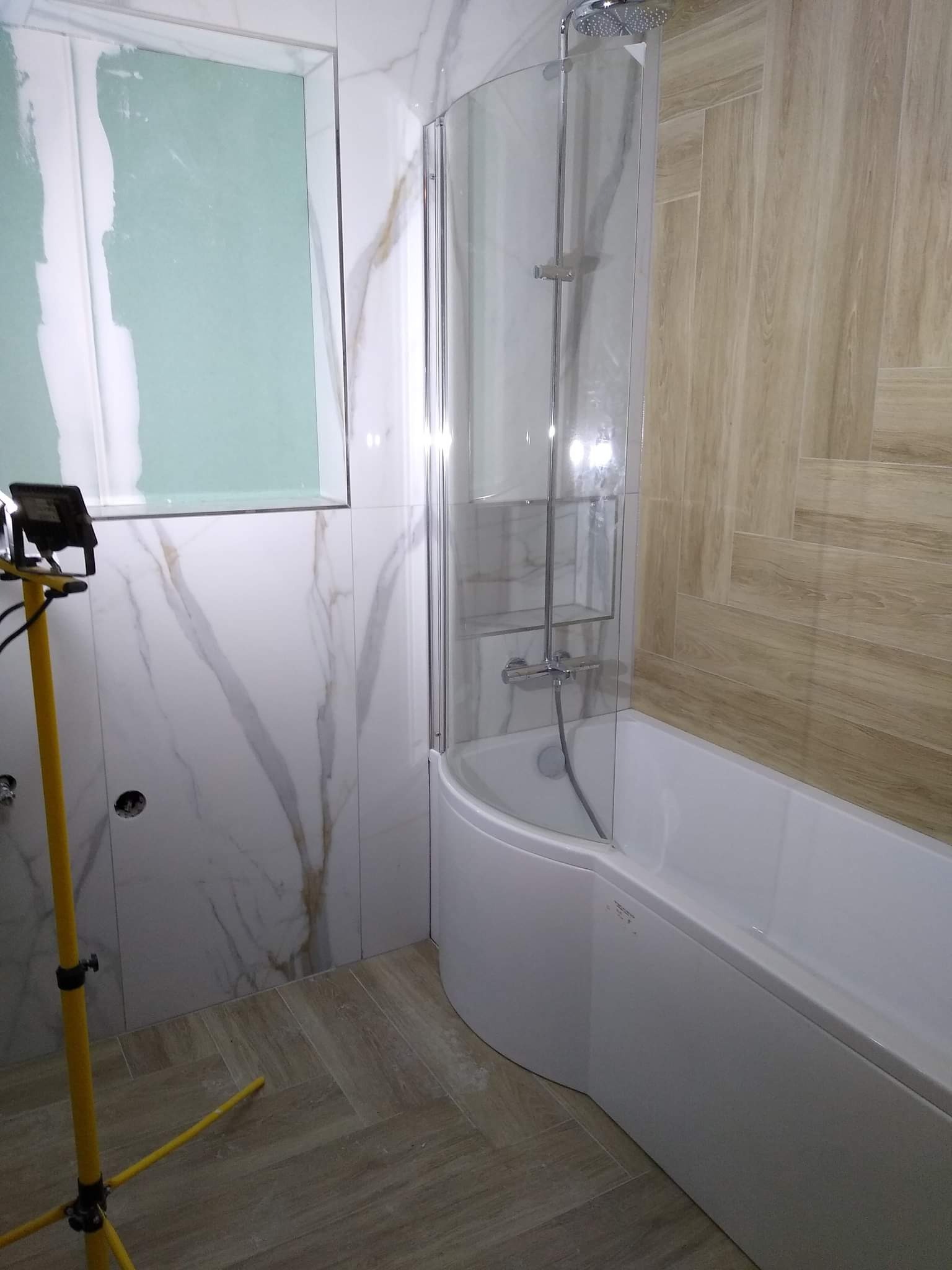 Niedokończona łazienka z wanną, szklanym parawanem nawannowym, marmurowymi płytkami ściennymi i podświetloną płytą kartonowo-gipsową nad wanną, oświetlana żółtą lampą budowlaną.