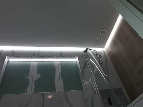 Nowoczesna łazienka z podświetlanym sufitem LED, marmurowymi płytkami i szklaną kabiną prysznicową, z której leje się woda.