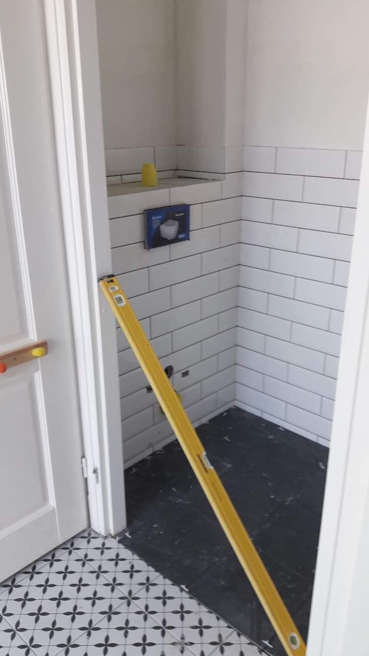 Wnętrze łazienki w trakcie remontu: białe, prostokątne płytki ułożone na ścianach z czarną fugą, czarna podłoga, poziomica oparta o futrynę drzwi, pudełko po stelażu WC na półce.